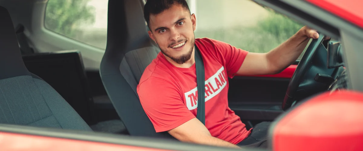 Führerschein Ratgeber: Ein junger Mann hält seinen Führerschein stolz in die Kamera, im Hintergrund ein Auto.